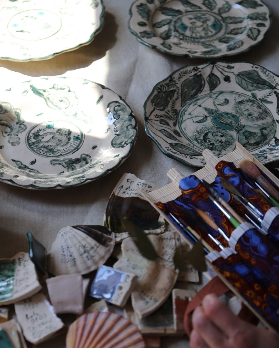 Lisa Sandner’s hand-painted plates aligned on a workspace table alongside fine-tip paintbrushes and ceramic pigments.