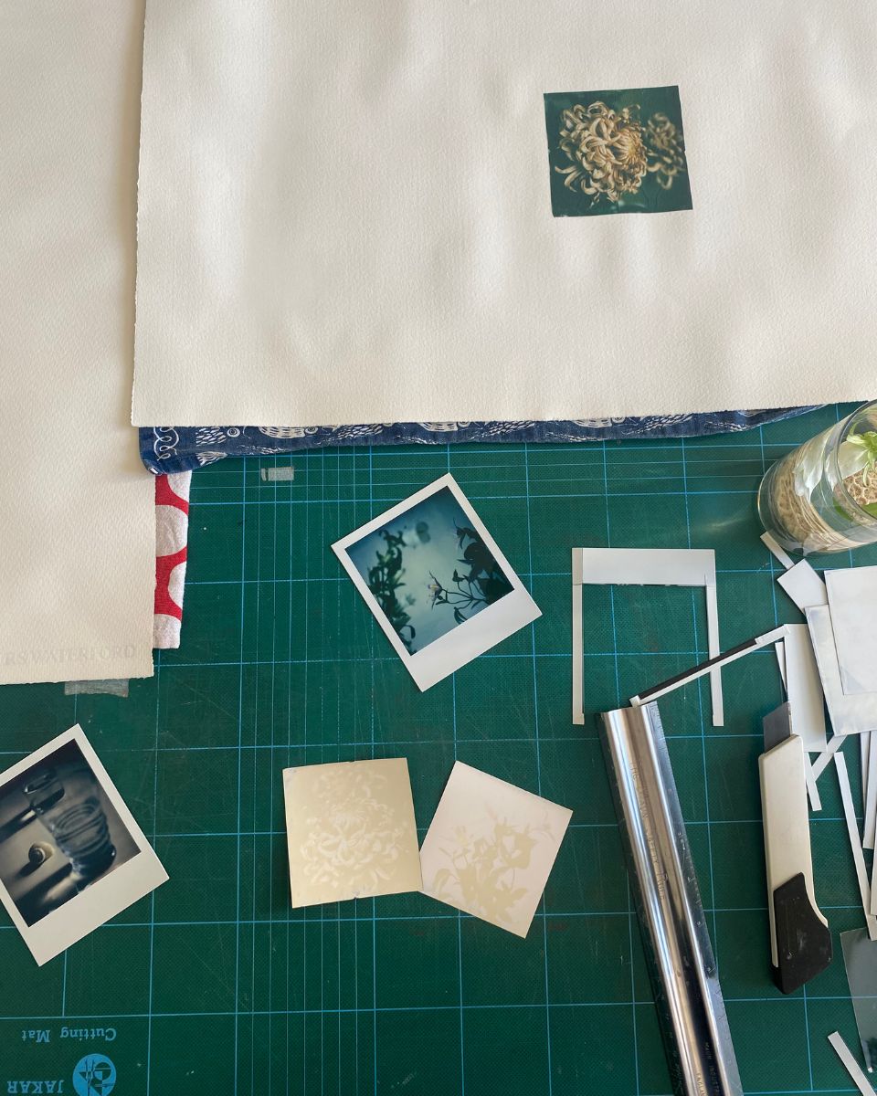 An artist's green cutting mat with craft tools, analogue Polaroids, and floral photography test prints in Lottie Hampson's studio