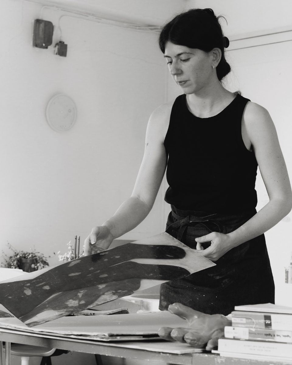 A black and white portrait of artist Mae Nicolaou at work inside her London studio space, featured for Partnership Editions.