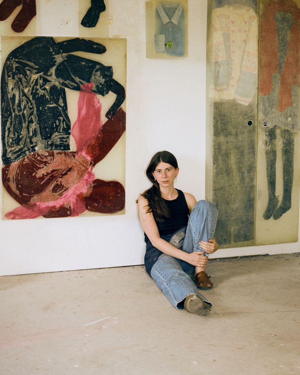 Seated portrait of artist Mae Nicolaou in front of a large sculptural print piece mounted on her wall.