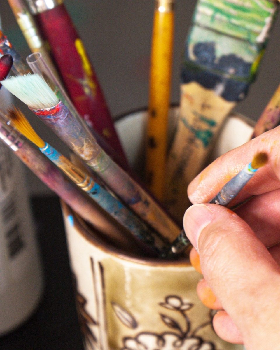 Macro detail of Mamie Young selecting a fine-tip brush from her studio pot, emphasising the delicate technique required for her smooth, air-filled forms.