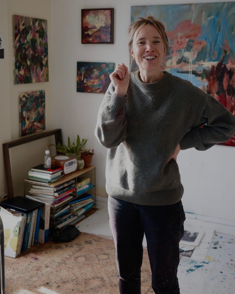 A smiling portrait of artist Mary West gesturing in front of abstract paintings hanging on her studio wall.