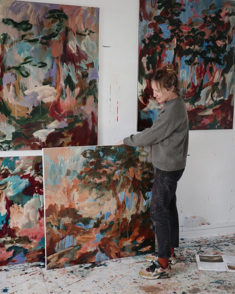 Full-body portrait of artist Mary West in her London studio, holding an original oil painting amidst a backdrop of large-scale abstractions.