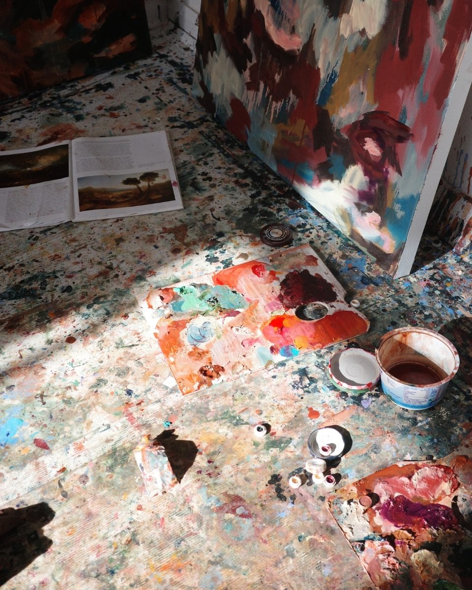 Overhead detail of Mary West’s paint-splattered studio floor, featuring used palettes, reference imagery, and open oil tubes.