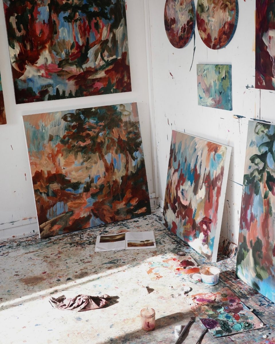 A corner view of Mary West’s studio workspace, displaying a curated collection of mounted and unmounted paintings on canvas.