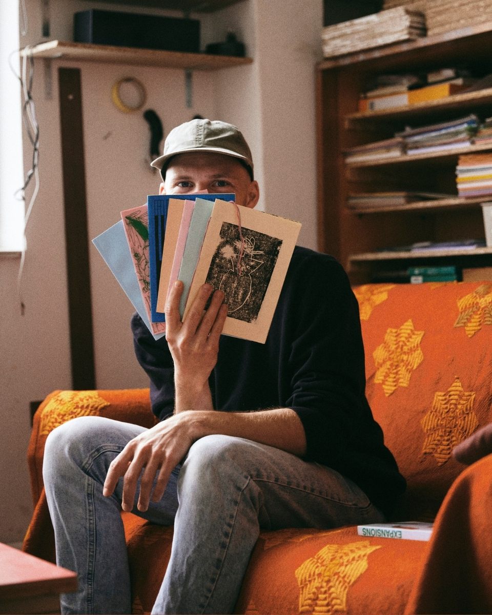 Artist Max Freund on an orange sofa, holding small colourful artwork prints in front of his face in his studio.
