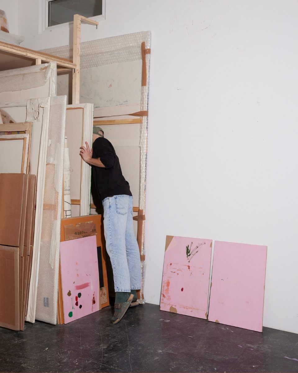 Full-length shot of artist Max Freund peaking playfully from behind a wooden storage rack filled with his contemporary slotted paintings.