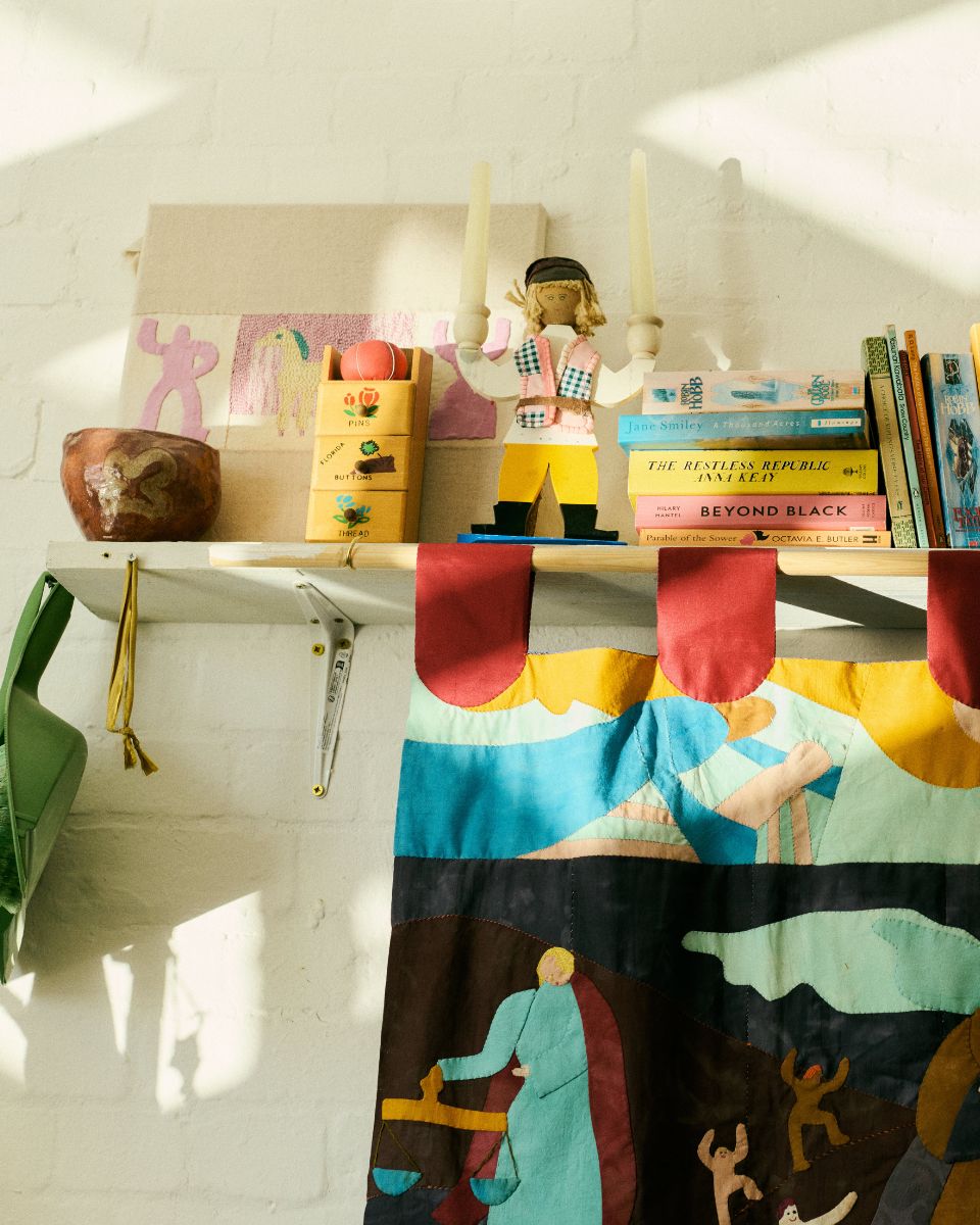 A styled wooden studio shelf holding colourful textile works, threads, reference books and small figurine.