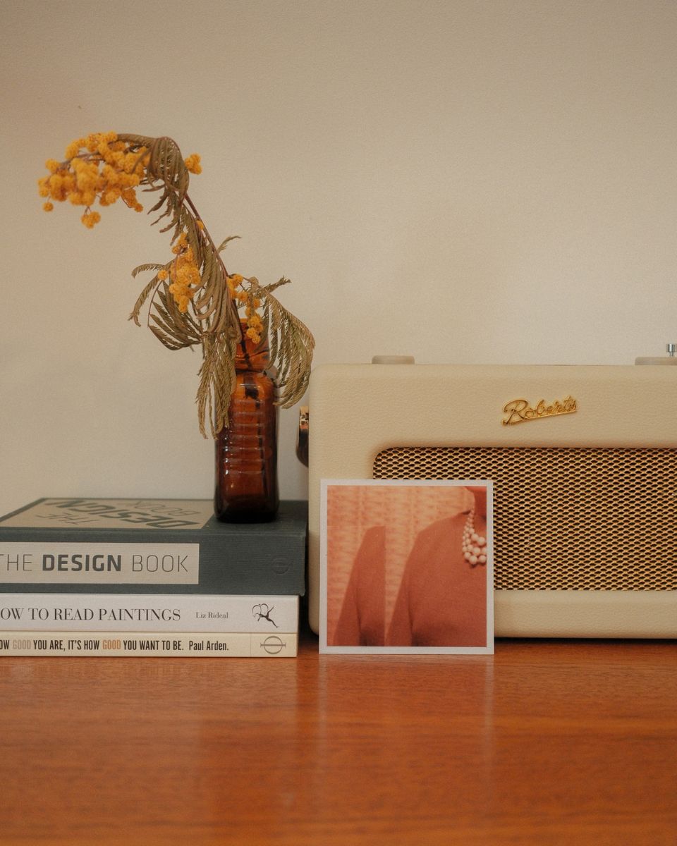 Dried yellow mimosa flowers in a glass bottle on design books next to a Roberts radio and portrait print.