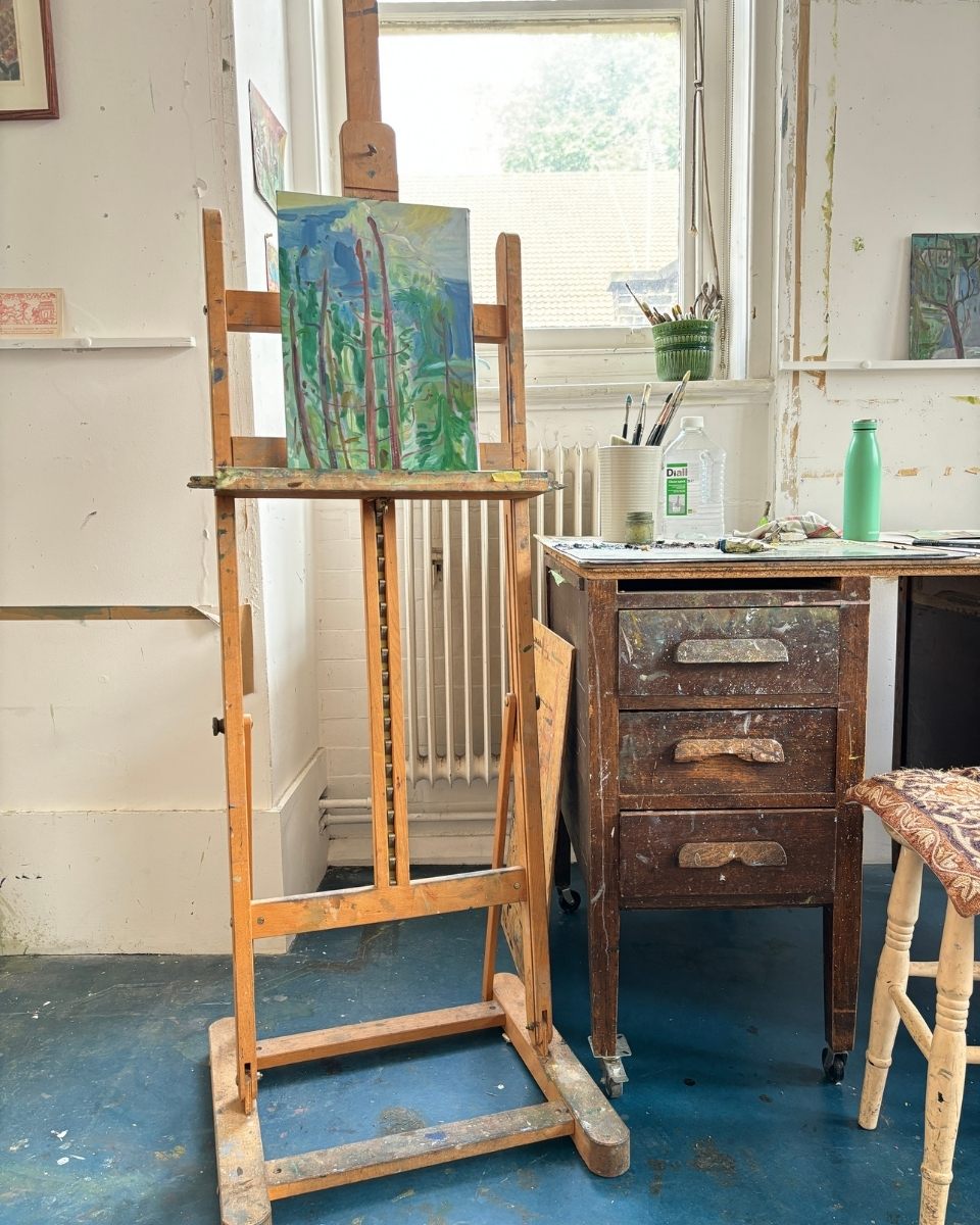 Detail of Nikki Gardham’s studio, featuring an atmospheric landscape on an easel and used paintbrushes on a paint-splashed wooden desk.