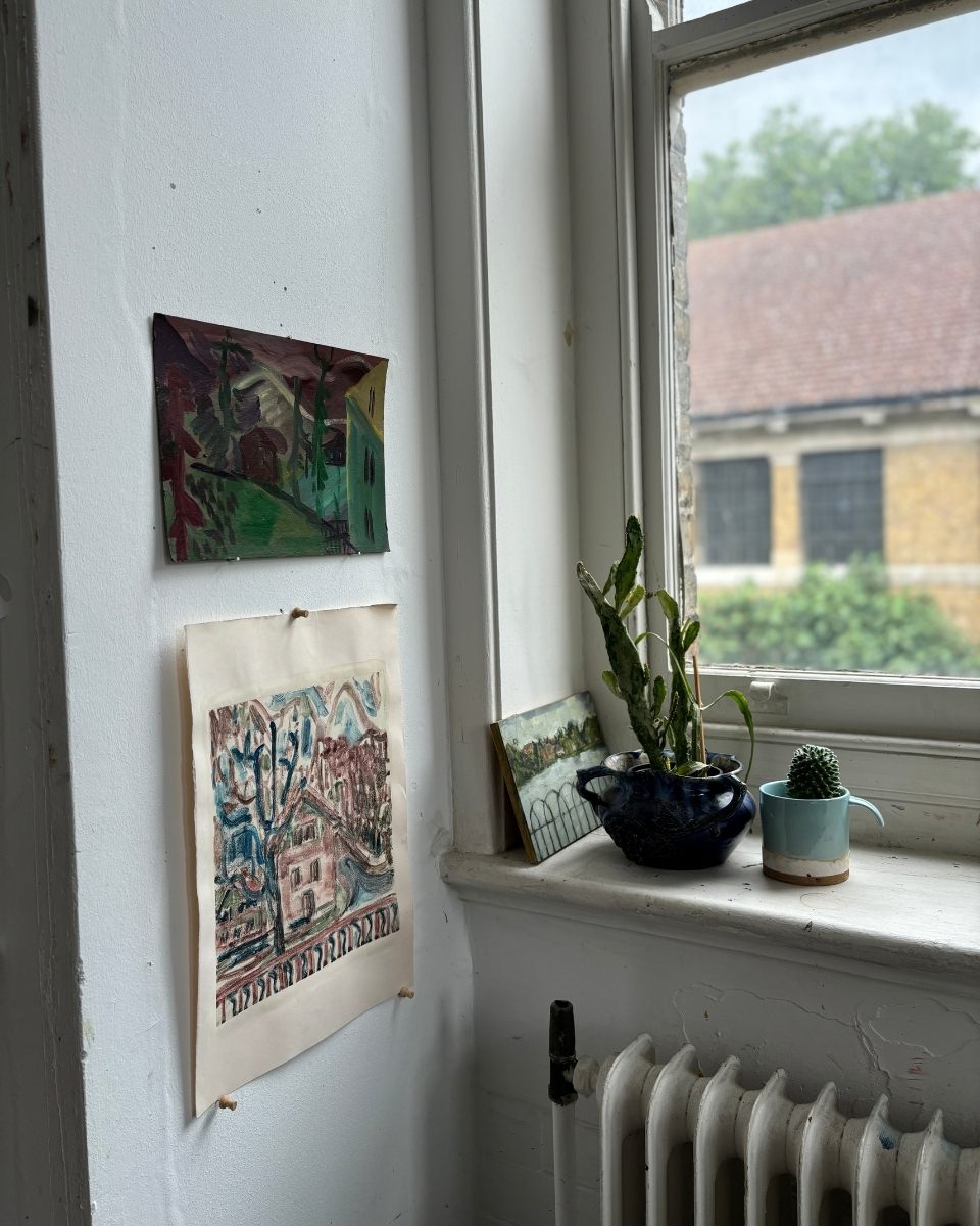 A studio corner by a window sill featuring small potted plants and sketch-like drawings pinned to the wall.