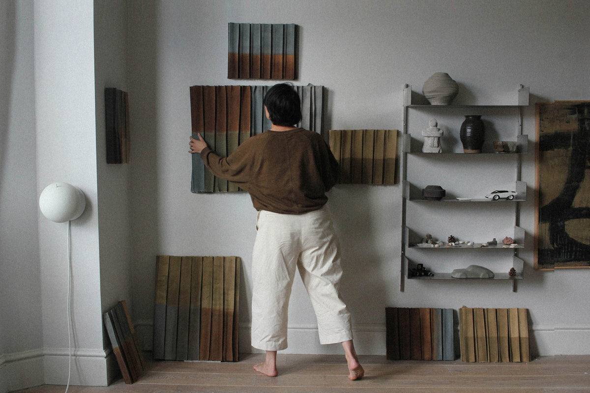 Textile artist Nikki Heaton arranging pleated earthy-toned fabric wall pieces in her minimalist studio workspace.