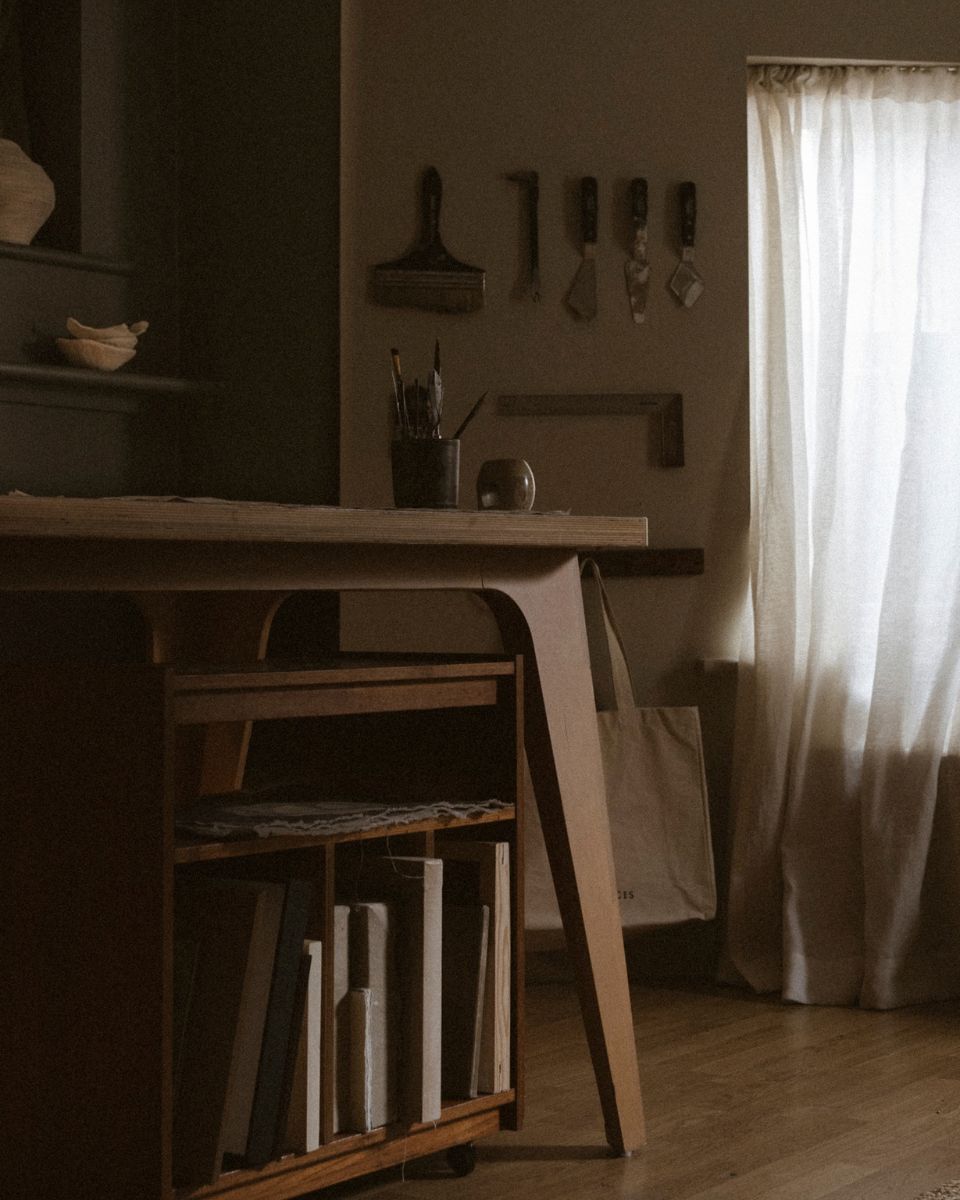 Warm wooden studio table with stored canvases beneath, palette knives on the wall, and linen sheer curtains.