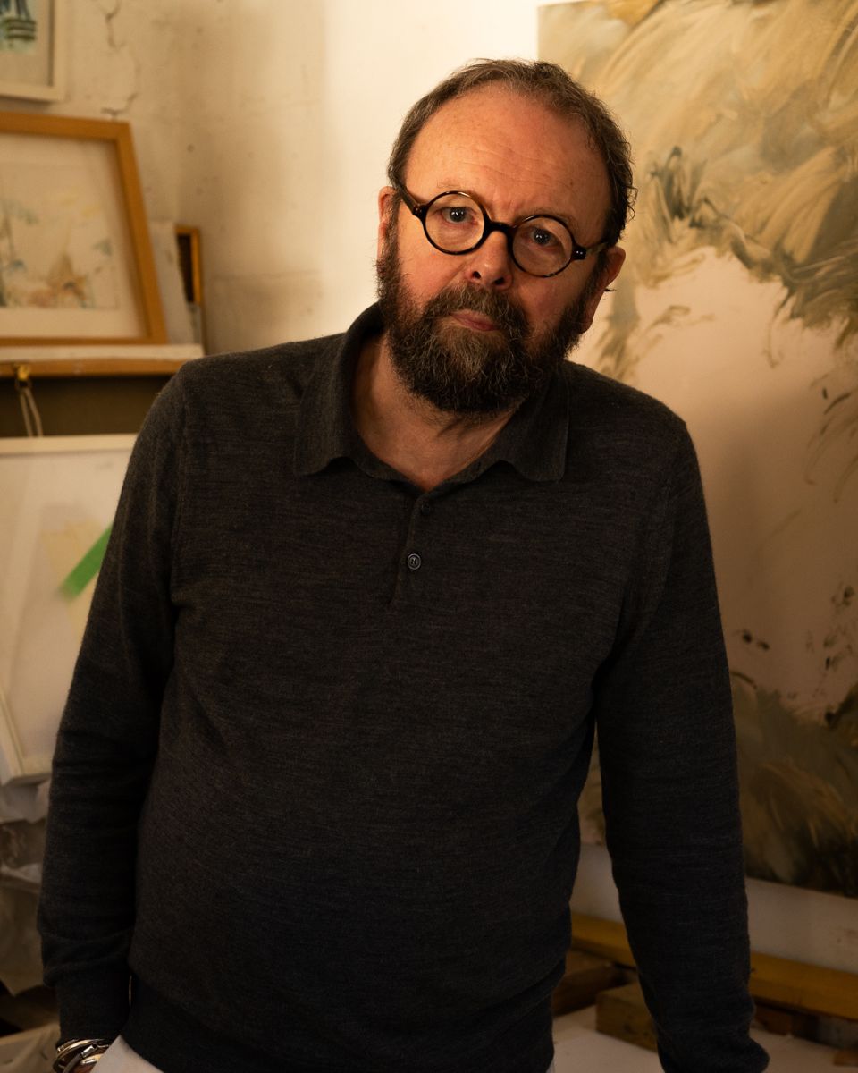 Artist Philip Maltman portrait, leaning on a table inside his studio surrounded by stacked canvases.