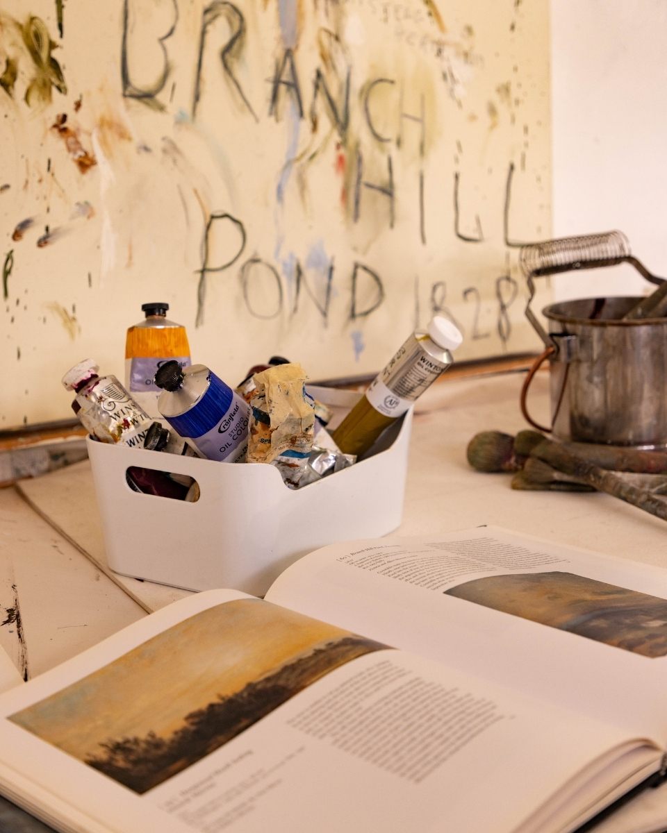 Detail of Philip Maltman’s studio featuring an open historical reference book alongside thick tubes of professional oil paints.