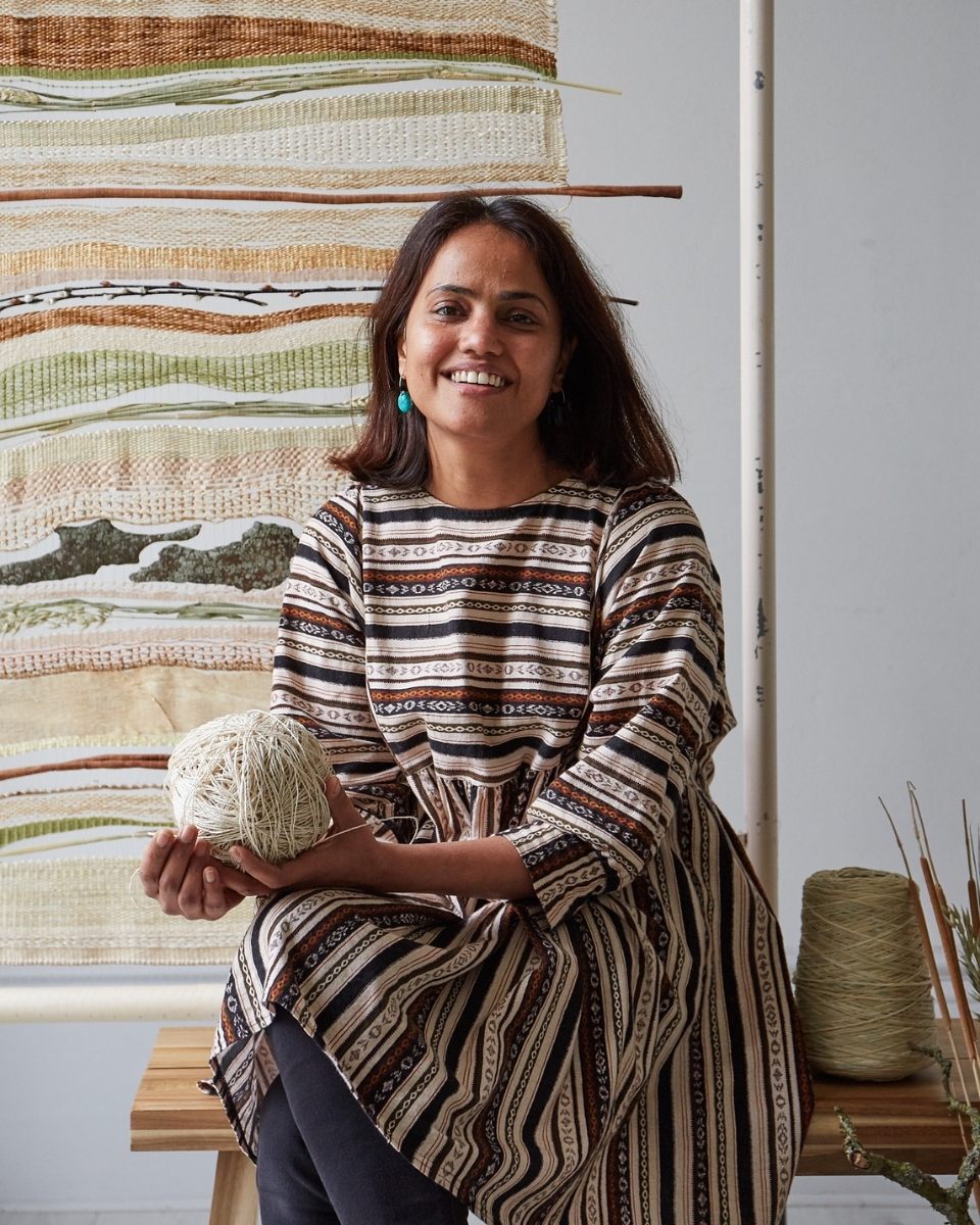 A smiling portrait of artist Rachna Garodia seated with a cotton handloom in her artist studio.