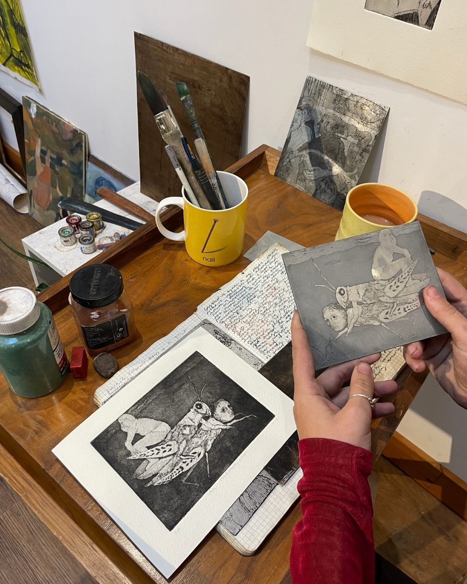 Rebecca Ashton in her studio holding a metal print card, showcasing the finish of her printed editions on a workspace table.