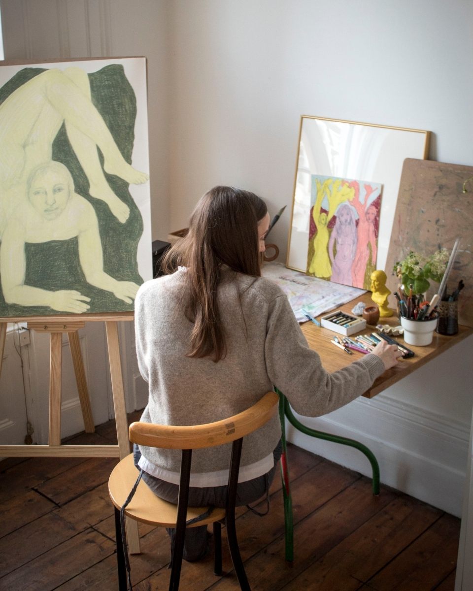 Artist Rebecca Sammon seated at her studio desk, focused on a vibrant composition on an easel.
