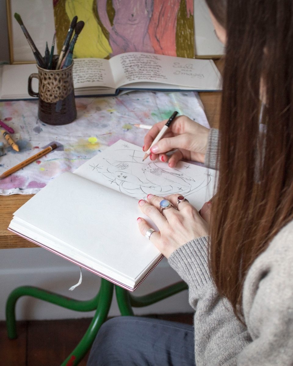 Close-up of Rebecca Sammon’s hands drawing in her sketchbook, capturing the gestural beginnings of her final compositions.