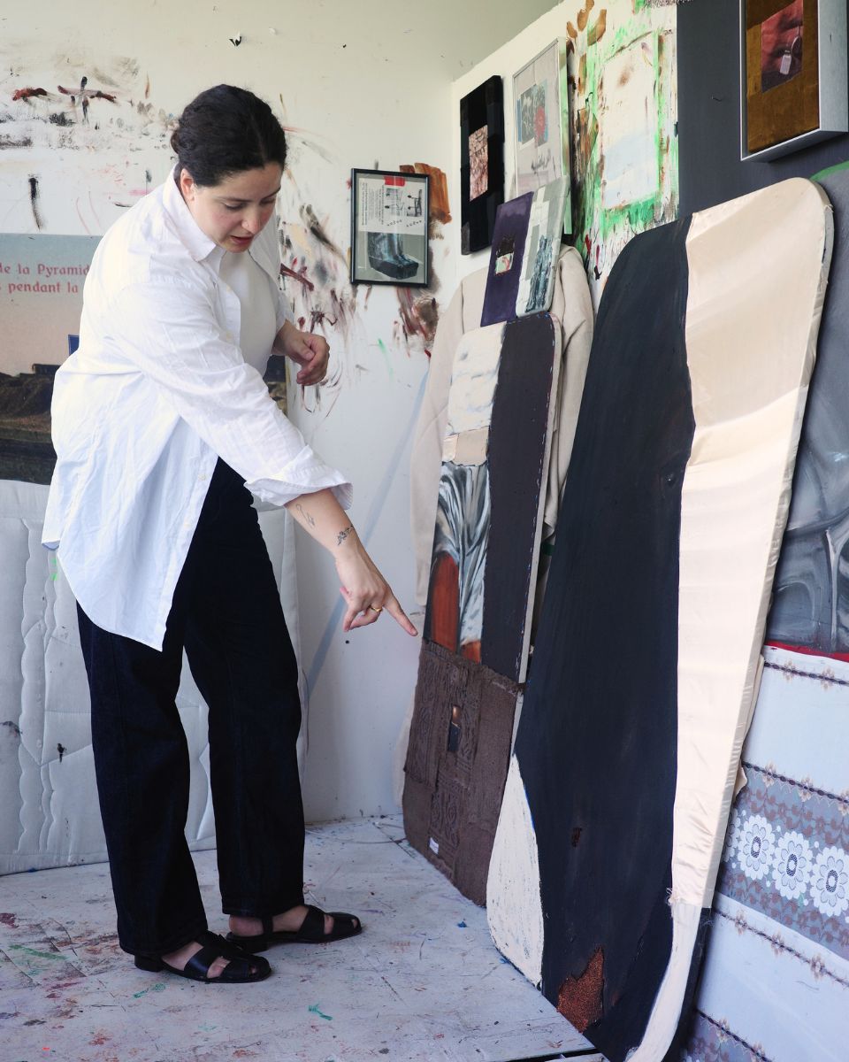 Artist Ruba Nadar pointing at a textured medium-scale mixed-media artwork resting on her studio floor.
