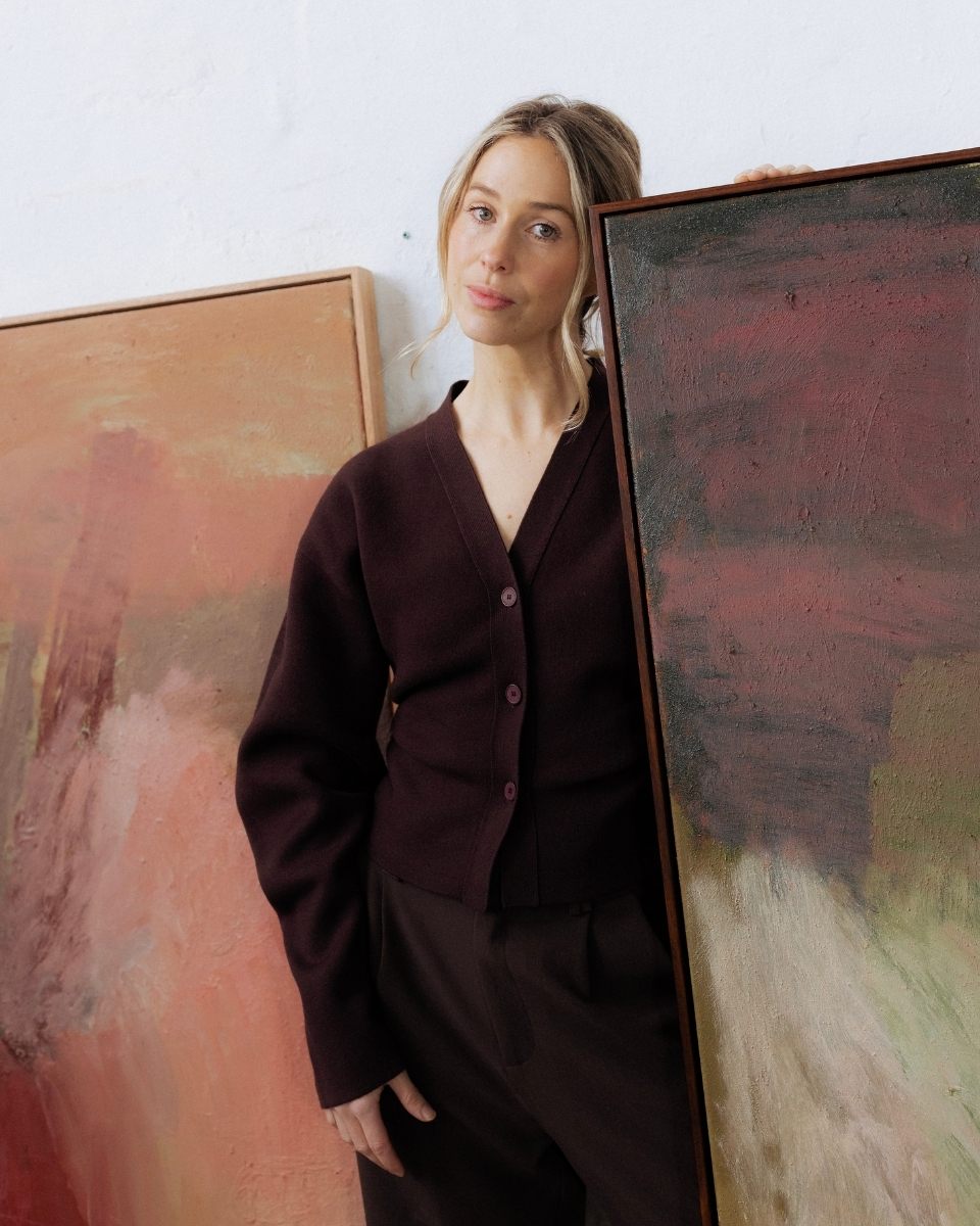 Sara Winfield framed by large abstract paintings, holding a canvas to her face in her art studio.