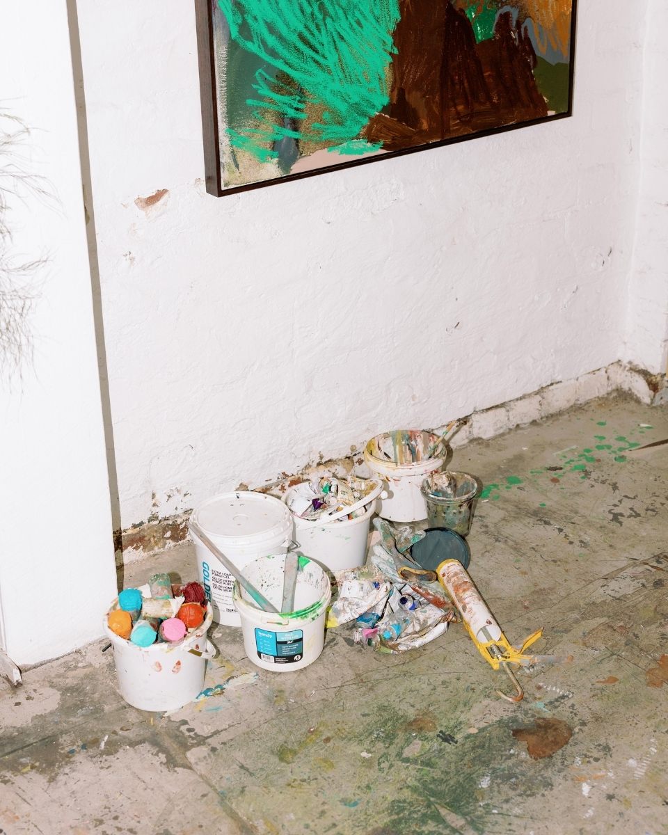 A top-down carousel image of Sara Winfield’s paint-splattered studio floor in Fremantle, scattered with paint tubs and a glimpse of a mounted abstract canvas.