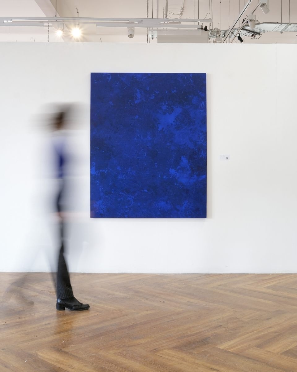 A slow shutter speed image capturing a viewer walking past a Simon Job painting in a gallery setting.