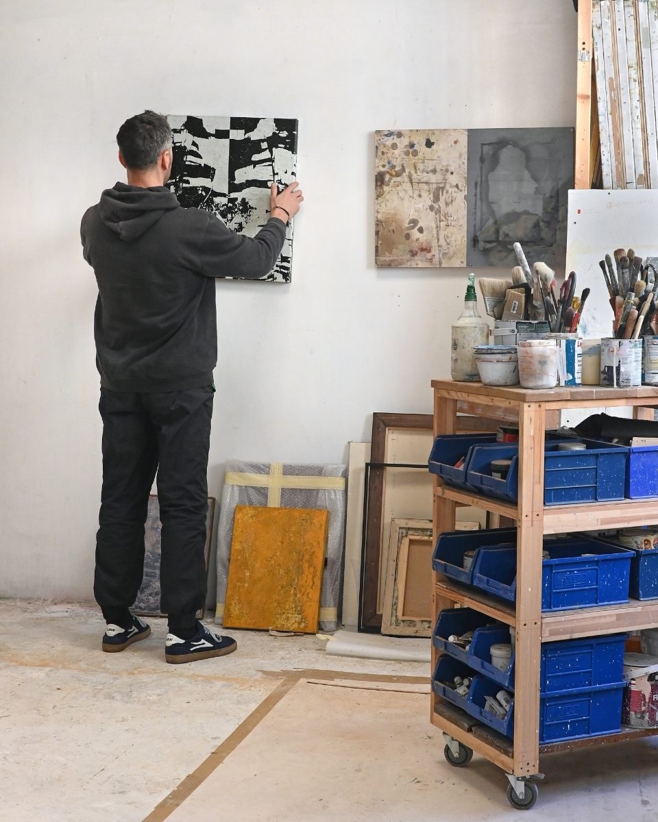 Simon Job arranging a painting on his studio wall, surrounded by his industrial tool trolley and other artworks.