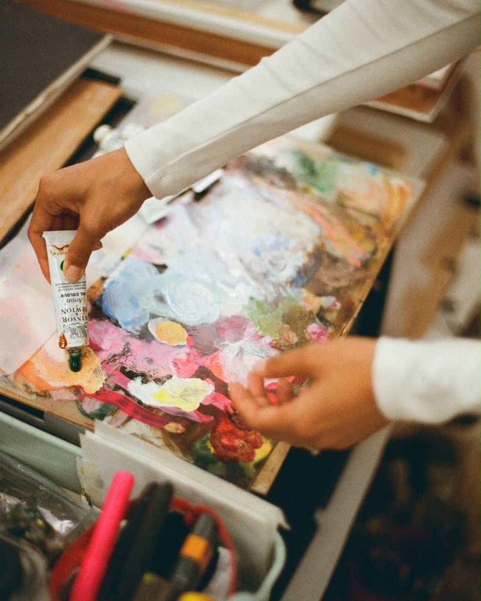 Artist hands squeezing Winsor & Newton oil paint tube onto a messy wooden palette board.