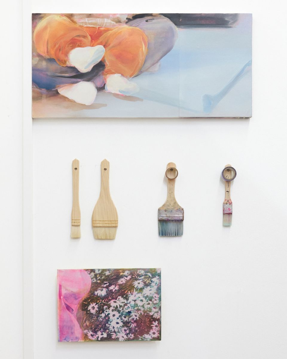 White studio wall displaying wide paintbrushes hung beneath a figurative painting and above a floral oil study.