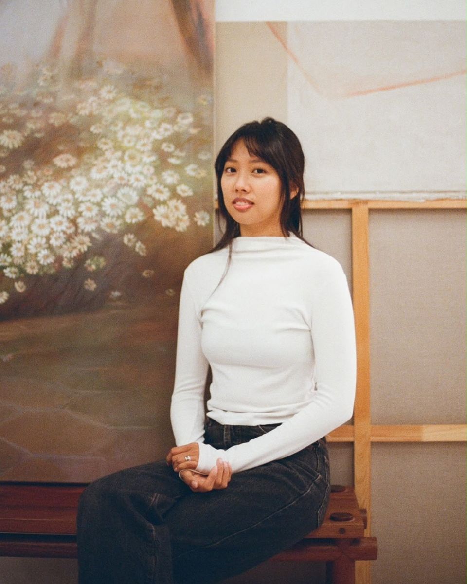 Portrait of artist Sophia Rosenthal wearing a white turtleneck sweater sitting in front of a daisy canvas in her studio.