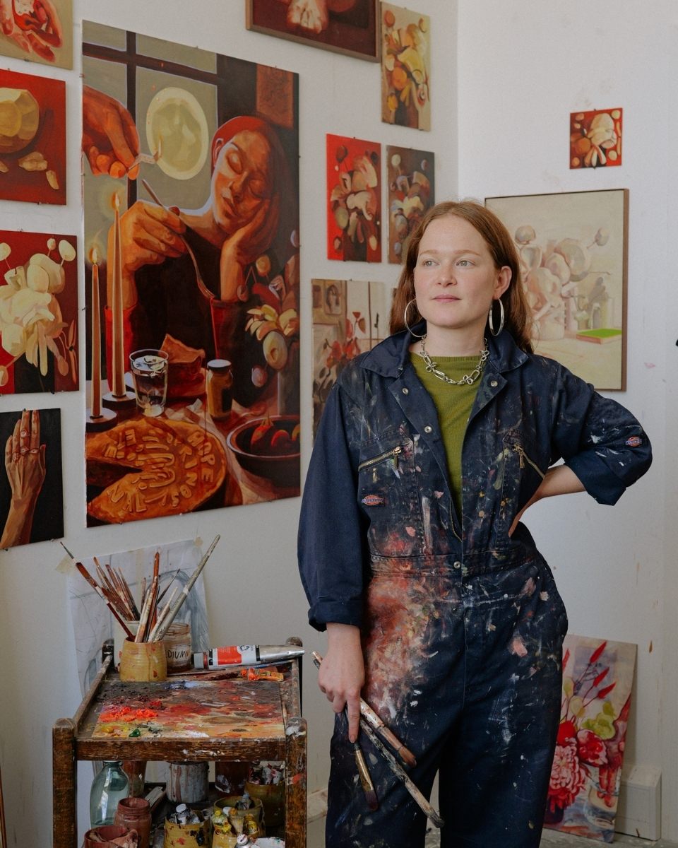 A serene portrait of Sophie Glover in her London studio, holding paintbrushes and surrounded by her expressive figurative and floral works.