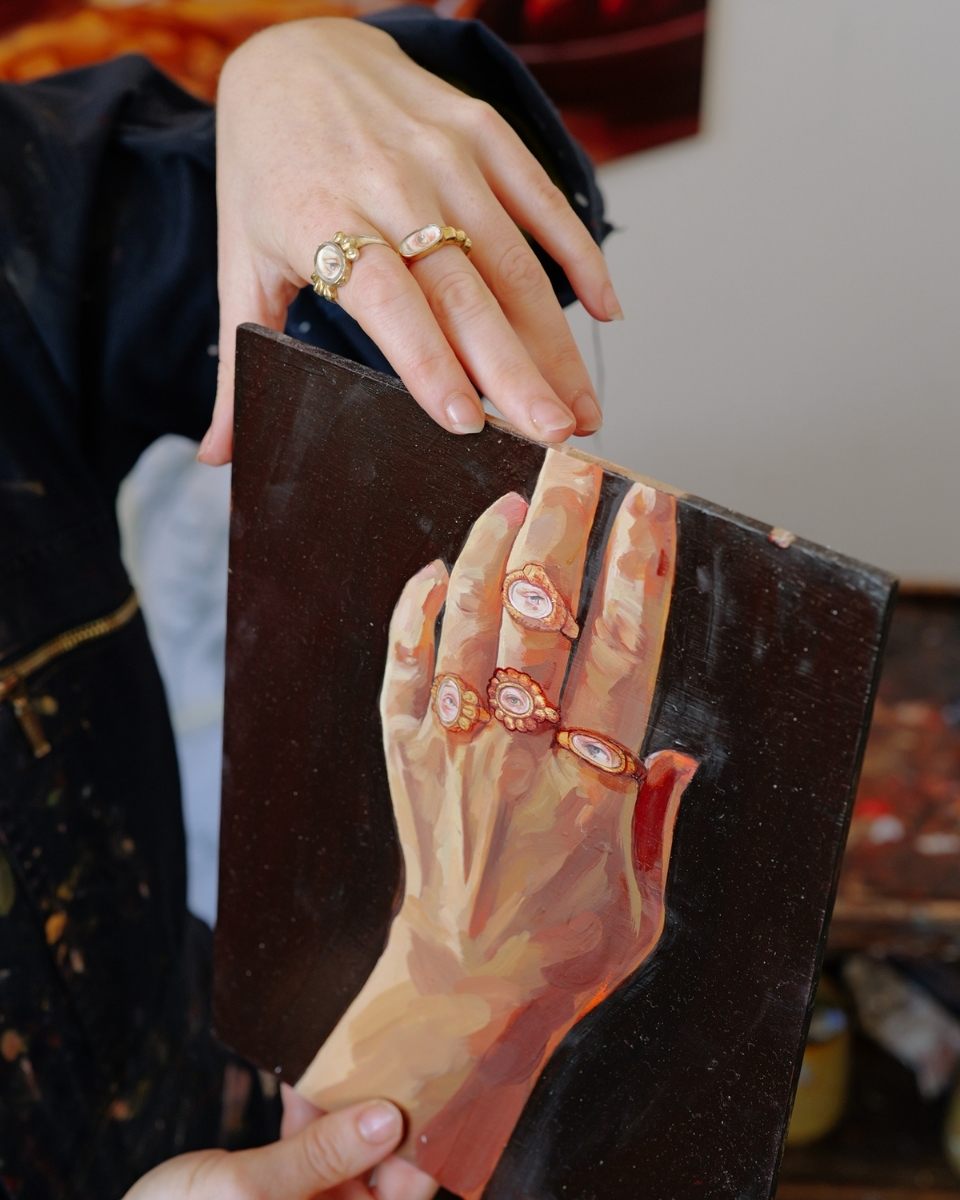 Sophie Glover wearing her hand-crafted rings while holding a painting of a hand wearing those same rings, bridging jewellery and fine art.
