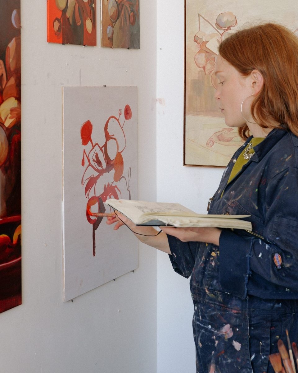 Detail of Sophie Glover painting a delicate floral composition on a wall-mounted wooden board while referencing her sketches in a handheld book.
