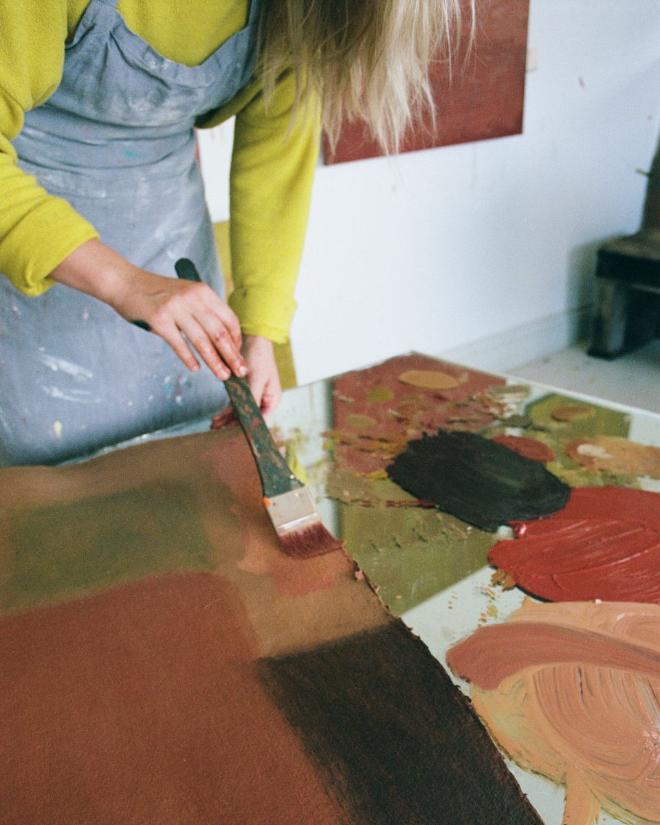 Close-up of artist Sophie Victoria Edwards applying a wide brushstroke of brown paint onto a textured canvas in her studio.