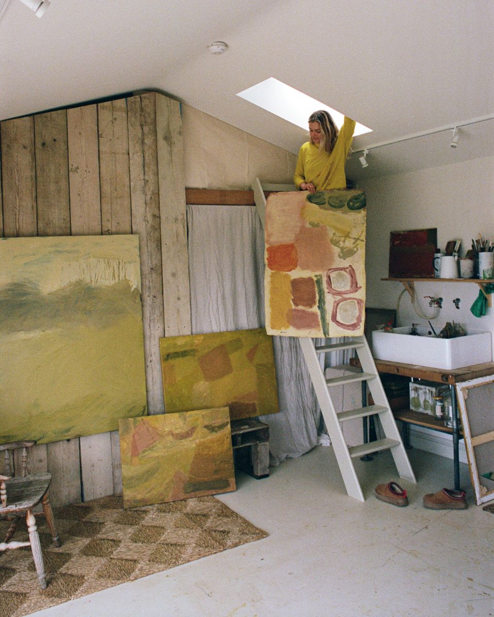 Wide view of artist Sophie Victoria Edwards standing on a studio ladder holding an abstract painting surrounded by her artwork and supplies.