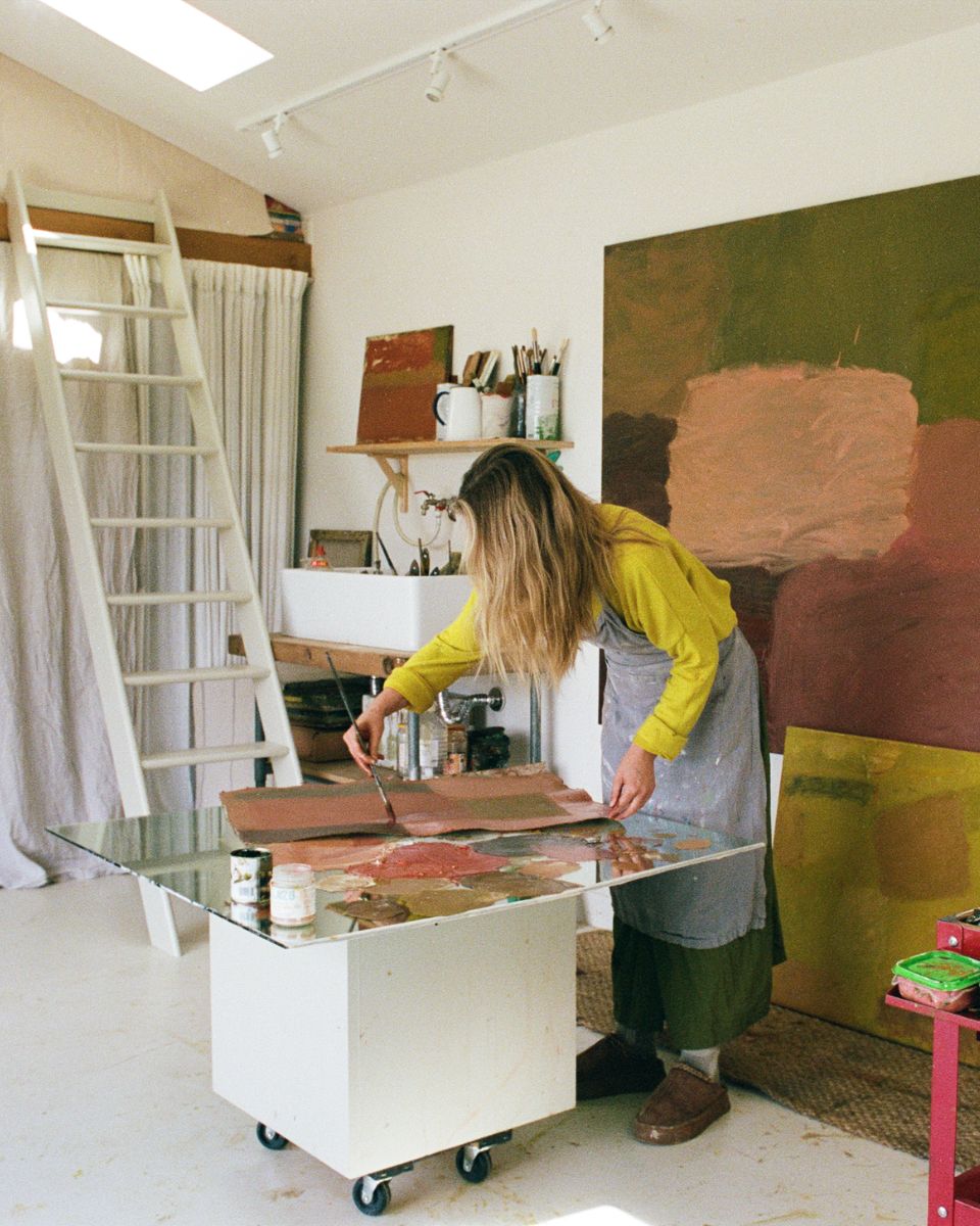 Artist Sophie Victoria Edwards leaning over a rolling work table with a glass surface, painting an artwork in her brightly lit studio.