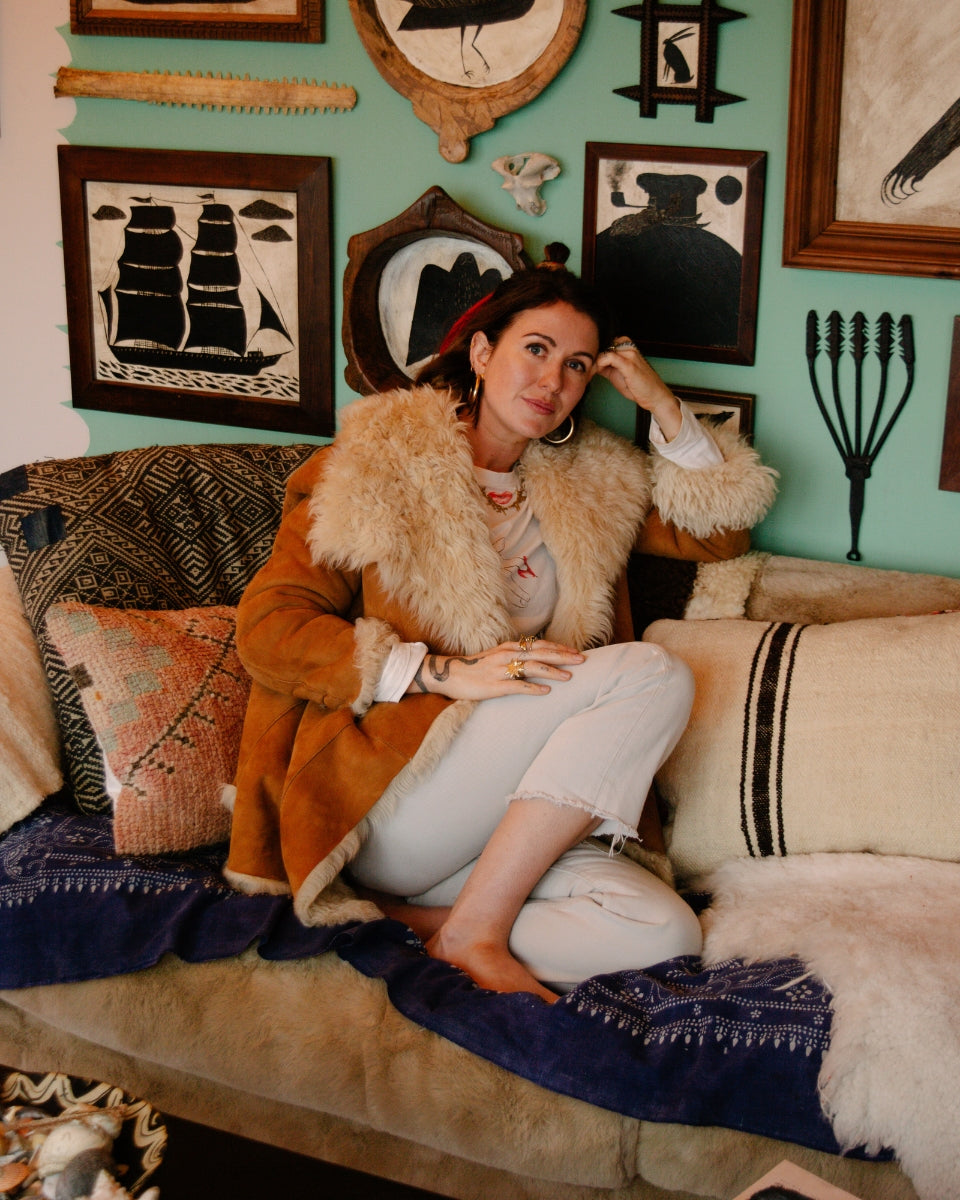 A seated portrait of artist Tabby Booth on a sofa in front of an eclectic gallery wall of her artwork.