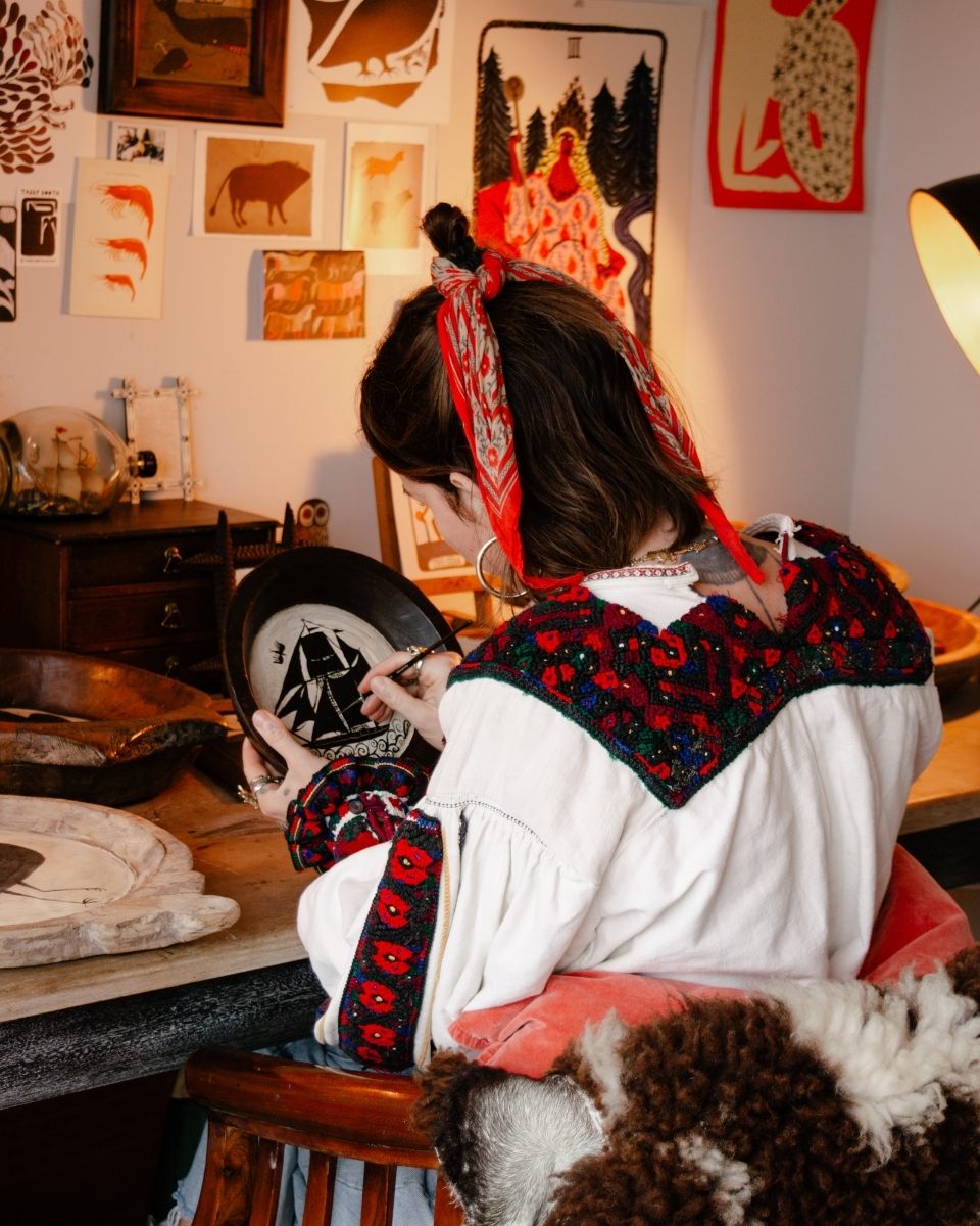 Sophie Booth shown from behind, meticulously painting a small-scale narrative artwork at her studio desk.