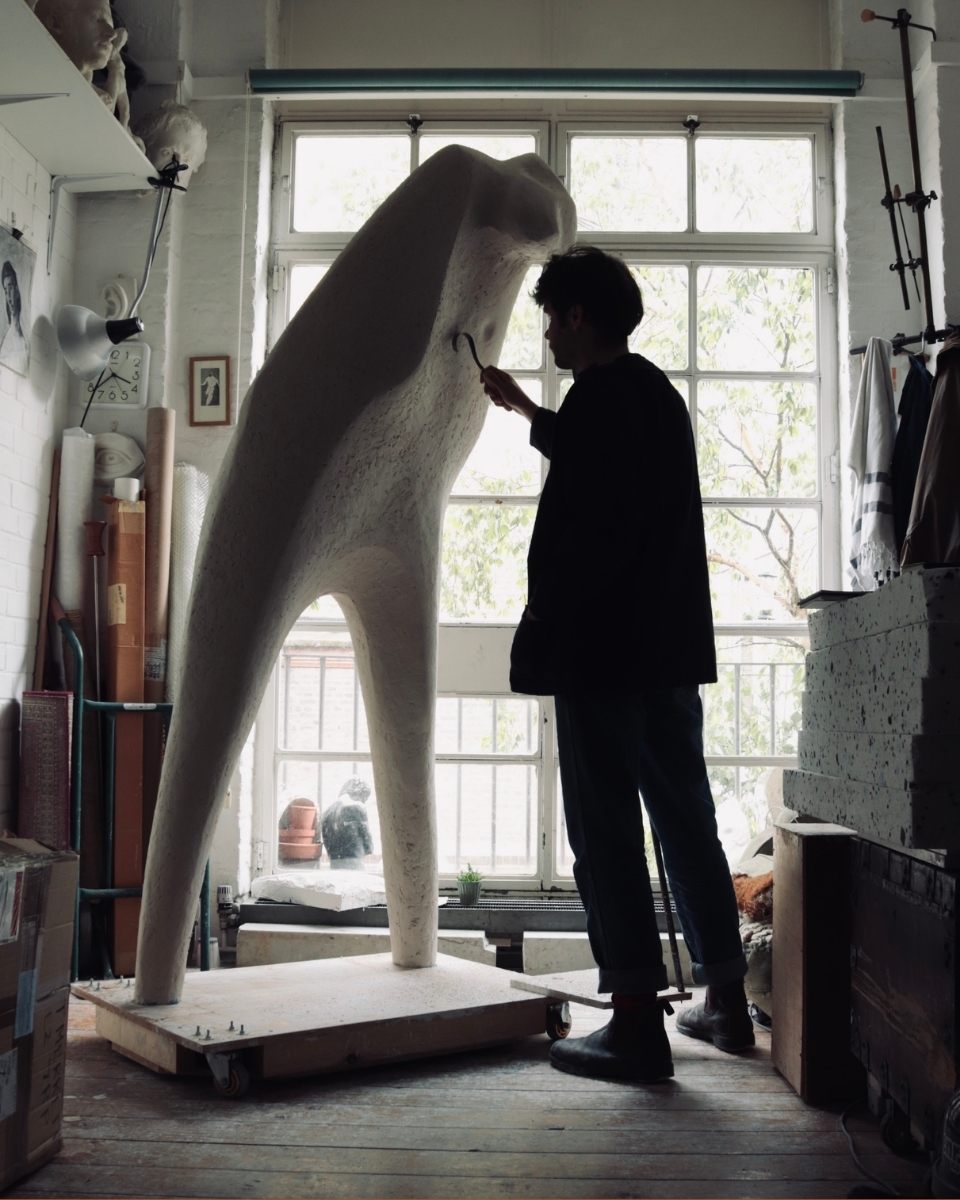 A moody, full-length shot of sculptor Thomas Merrett working on a large-scale human sculpture.