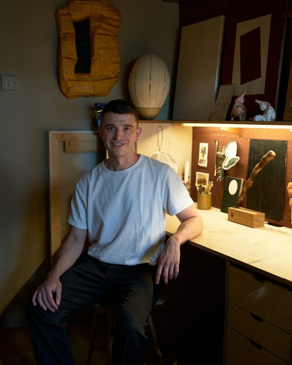 Portrait of artist Tom Collison at his studio desk in front of shelves holding his paper art and sculptures.
