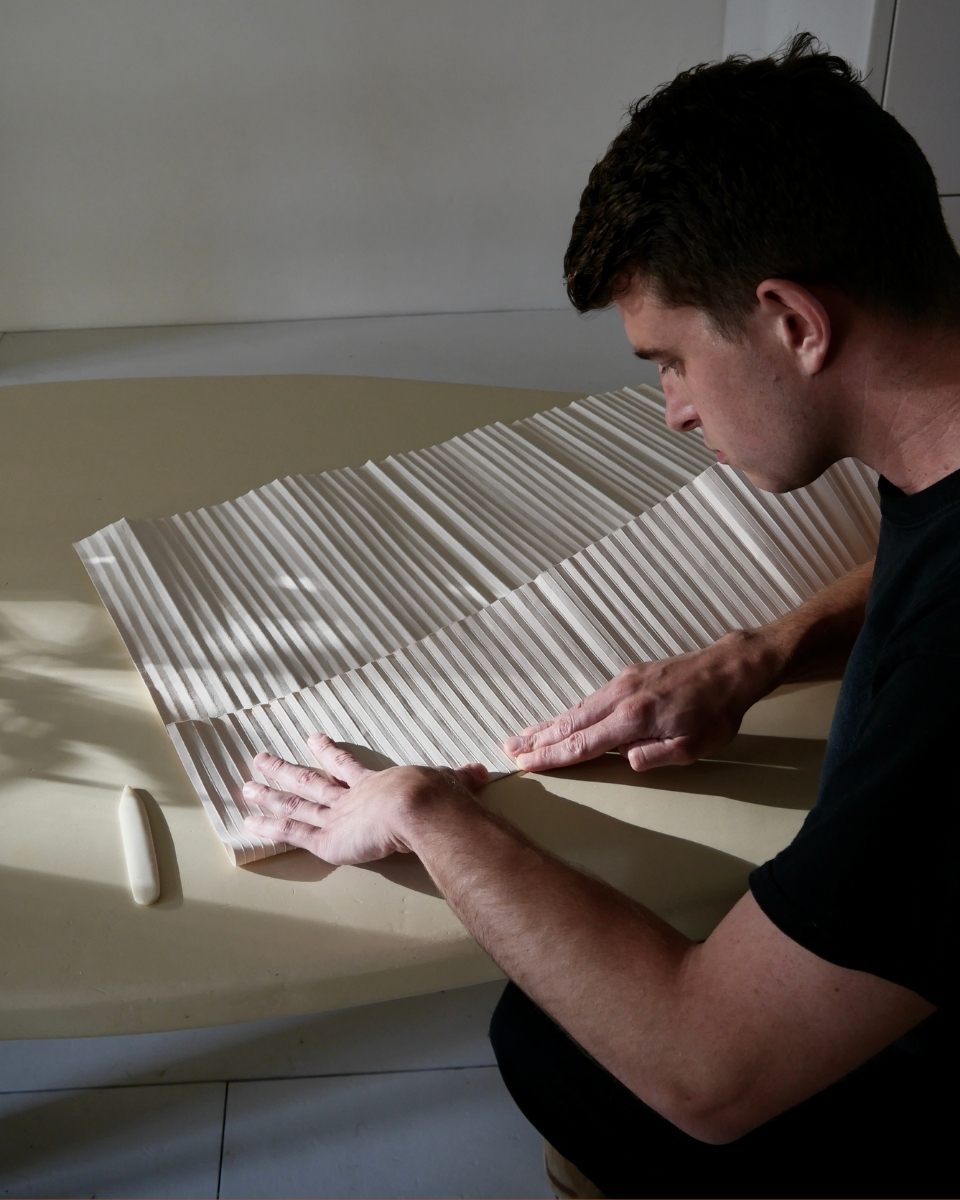 A process shot of Tom Collison’s hands meticulously folding mid-weight paper to create the rhythmic shadows of his relief artworks.