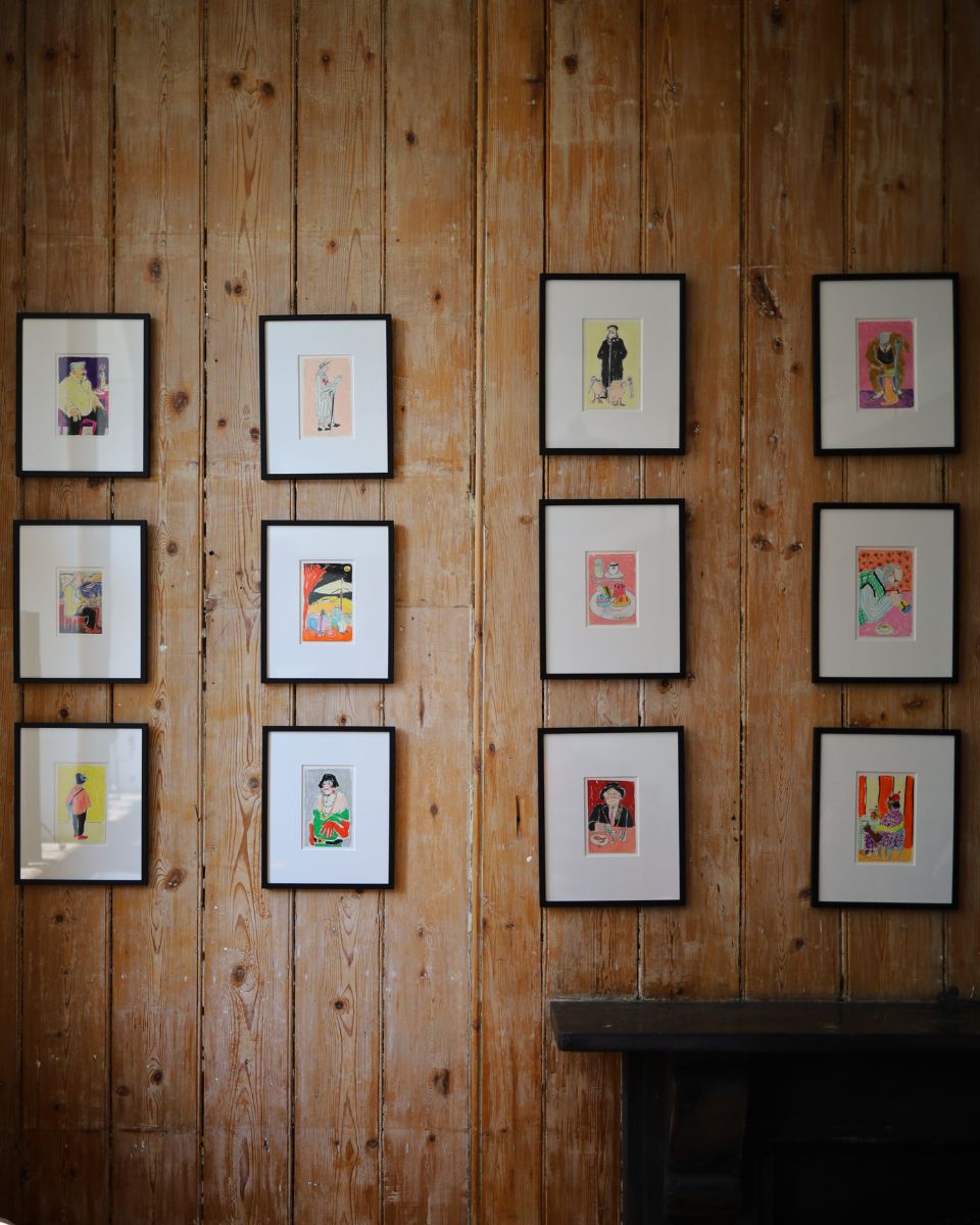 A collection of twelve framed, colourful original sketches by Isabella Cotier displayed in a grid pattern on a natural wood-paneled wall.